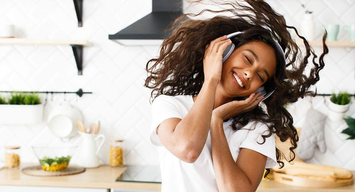Smiled mulatto girl with curly hair in big wireless headphones is happily dancing with her eyes closed in the modern kitchen